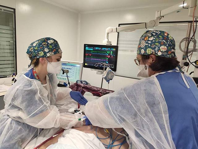 Un chirurgien en tenue stérile verte opère un patient allongé sur une table d'opération recouverte de champs chirurgicaux bleus dans un bloc opératoire équipé de divers appareils médicaux.